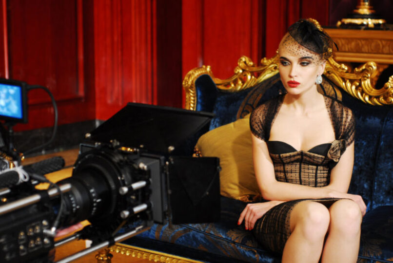 Woman sitting in front of a camera wearing a black dress and fascinator.