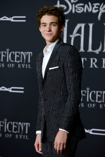 Joshua Bassett in a black with white striped tuxedo. He is smirking at the camera and this photo is taken place at the Disney Maleficent premiere.