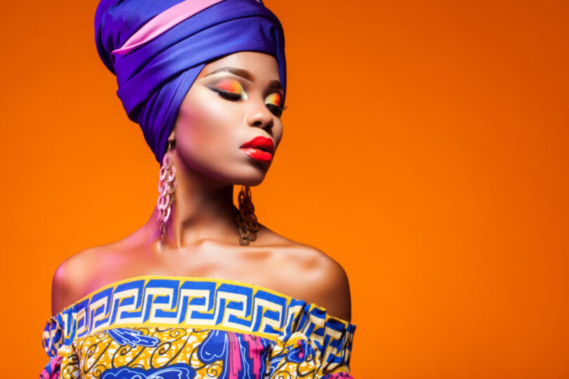 African woman in brightly colored dress and headwrap on orange background