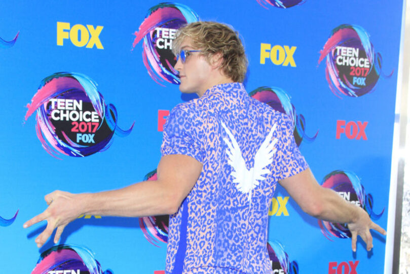Logan Paul at the Teen Choice awards wearing a purple shirt with a white maverick on his back. He is wearing sunglasses and giving us a side pose.