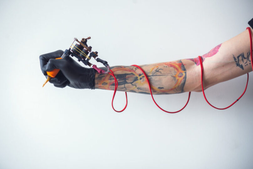 Guy holding tattoo gun with cord wrapped around his arm, only his one arm showing