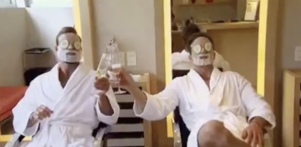 Two guys sitting in robes at a spa cheersing with champagne and cucumbers on their eyes