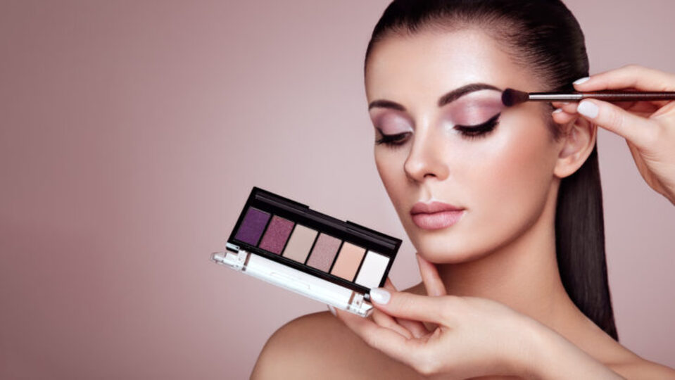 Makeup artist applying pinkish purple eyeshadow to beautiful young woman with dark hair and tan complexion