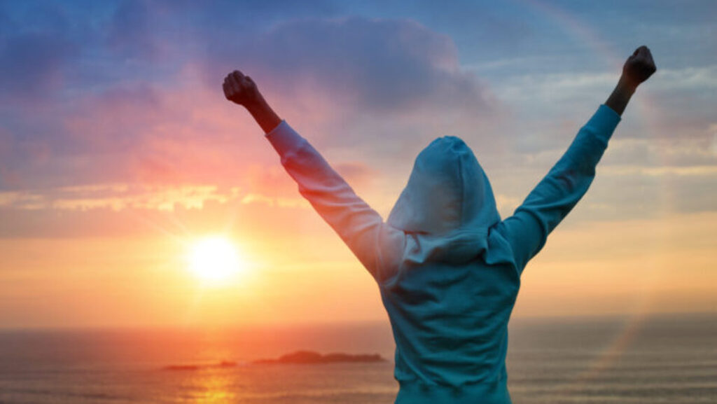 A woman looking out to the sunset with her arms up to celebrate her achievement 